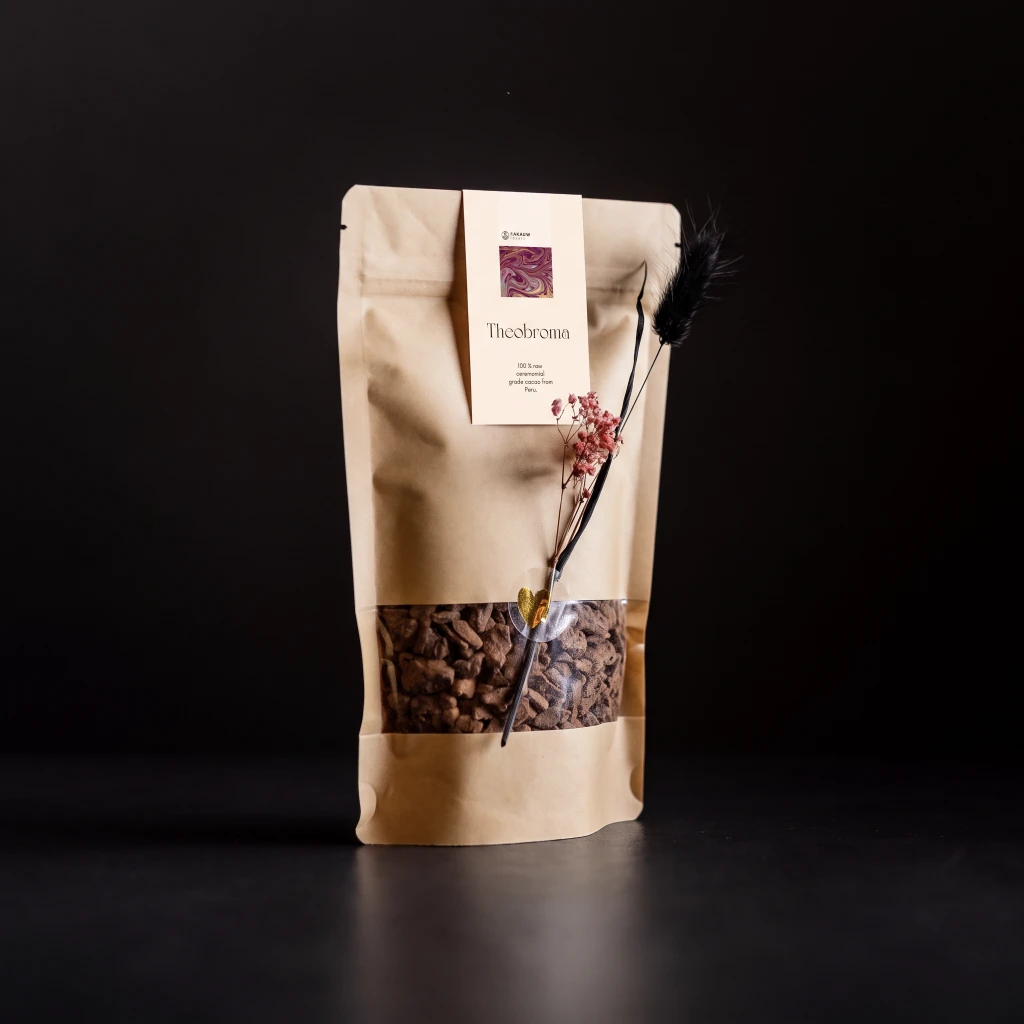 Kraftzak Theobroma ceremoniële cacao van Kakauwlovers met venster, gedroogde bloemen en gouden hartsticker op zwarte achtergrond